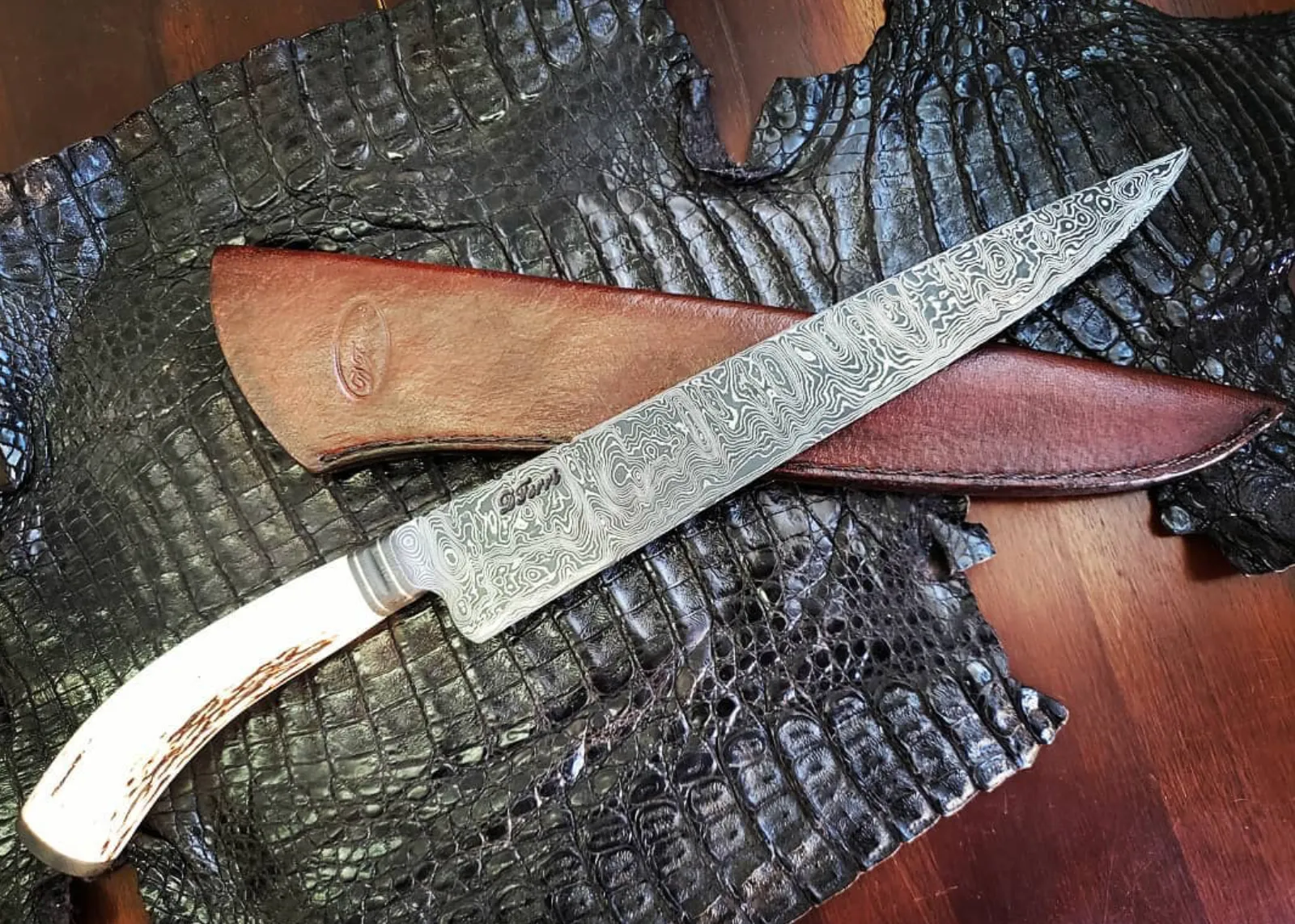 Damasco assinado DFerri, cabo em galha de veado e bainha de couro, sobre couro escuro escamado.